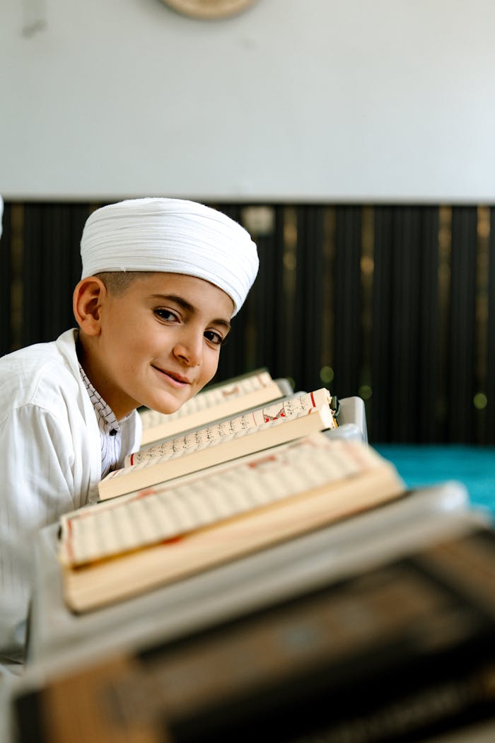 A young boy in traditional attire reads the Quran inside a mosque, smiling warmly.